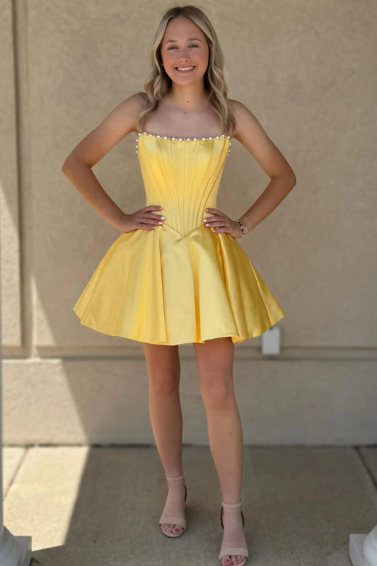 A woman modeling a short yellow strapless dress with a pleated corset-style bodice and flared skirt, standing against a beige stucco wall.