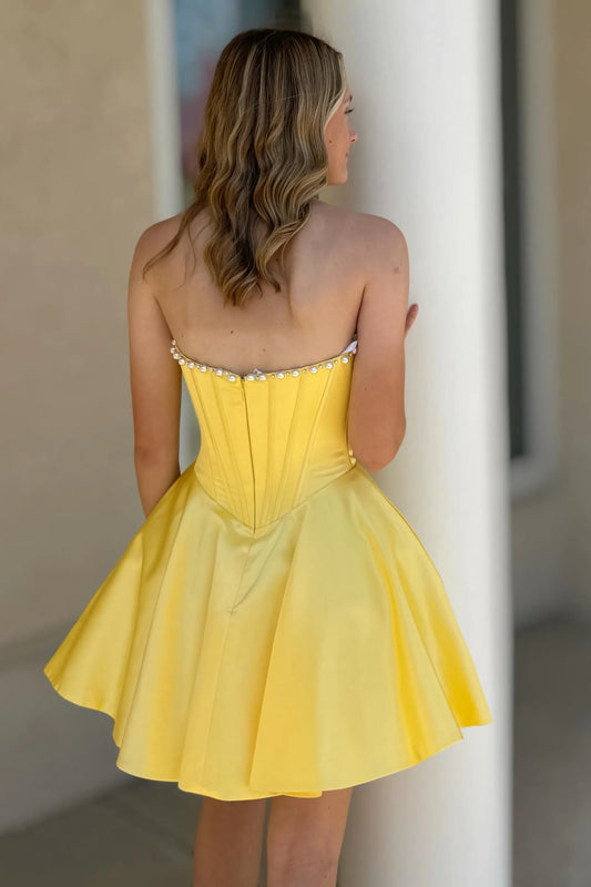 A woman wearing a yellow strapless mini dress, shown from the back as she looks to the side. The dress features a structured corset-style bodice with a flared skirt and delicate pearl embellishments along the straps.