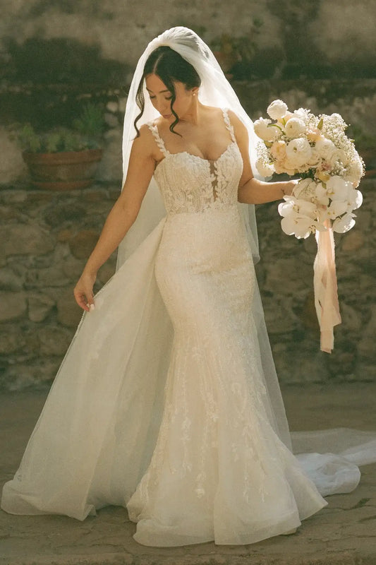 A bride in a fitted ivory lace wedding gown with a long sheer veil and a bouquet of white flowers, standing outdoors against a rustic stone wall.