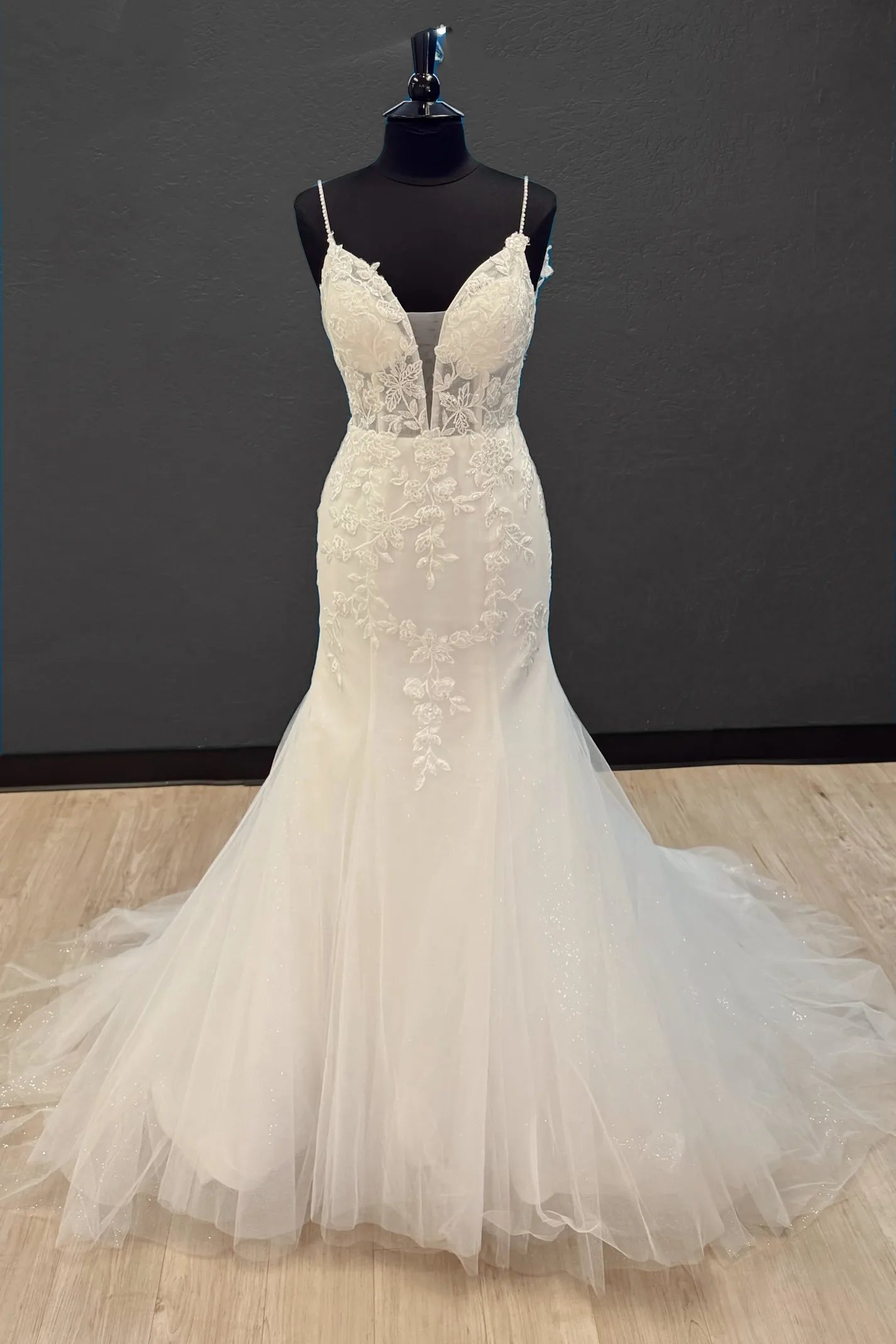 An ivory white lace and tulle wedding gown displayed on a black dress form mannequin against a dark gray wall and light wood floor.