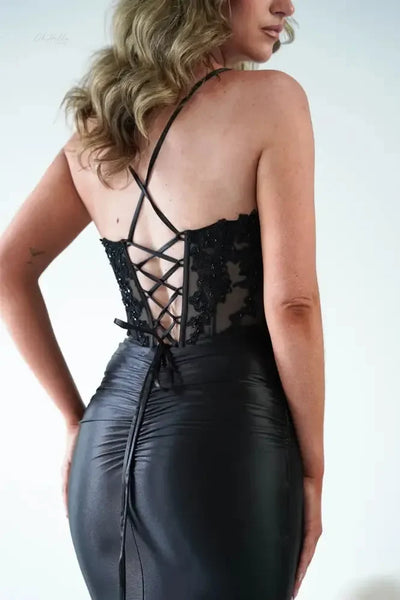 A woman modeling a floor-length black evening gown with a sheer lace bodice, thin spaghetti straps, and a fitted satin skirt, standing against a white backdrop with black draping.