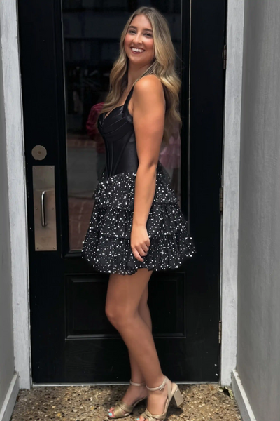 A woman modeling a short black dress with a structured corset-style bodice and a flared skirt covered in small white polka dots, standing in front of a black door.