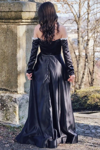 A woman wearing a long black evening gown with a sweetheart neckline, embellished with silver rhinestones, and featuring a high thigh slit, standing outdoors on a stone path.