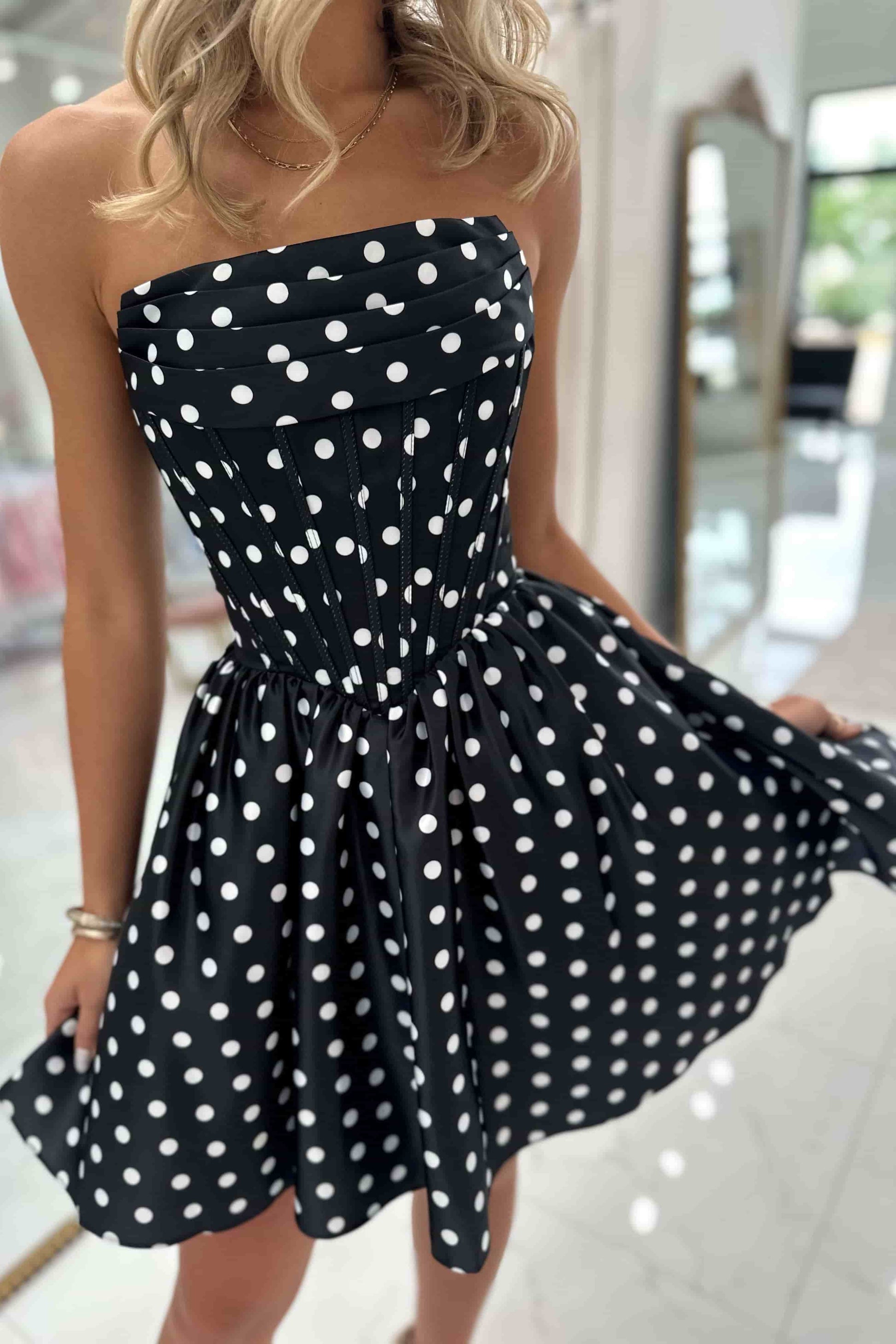 A woman wearing a strapless black dress with white polka dots, holding out the skirt to show its full flare in a bright indoor setting.