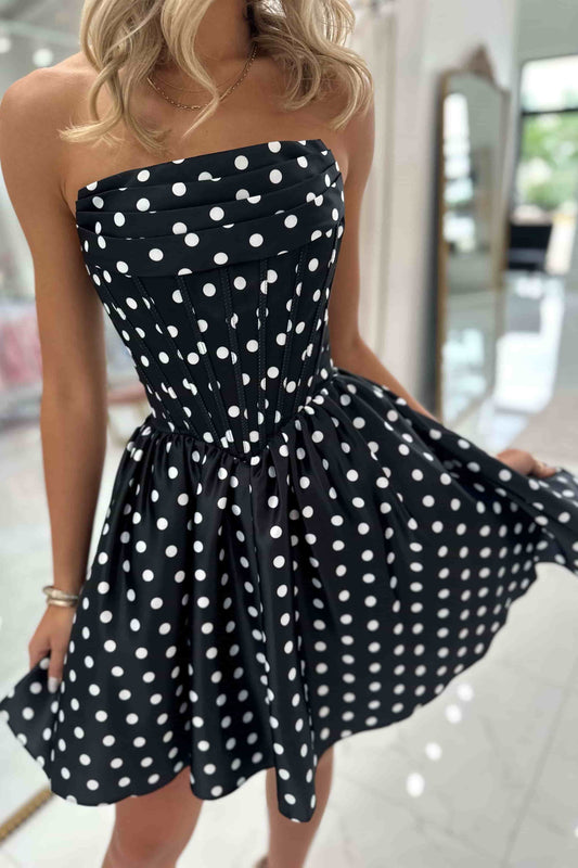 A woman wearing a strapless black dress with white polka dots, holding out the skirt to show its full flare in a bright indoor setting.