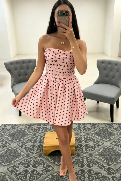 A woman wearing a strapless black dress with white polka dots, holding out the skirt to show its full flare in a bright indoor setting.