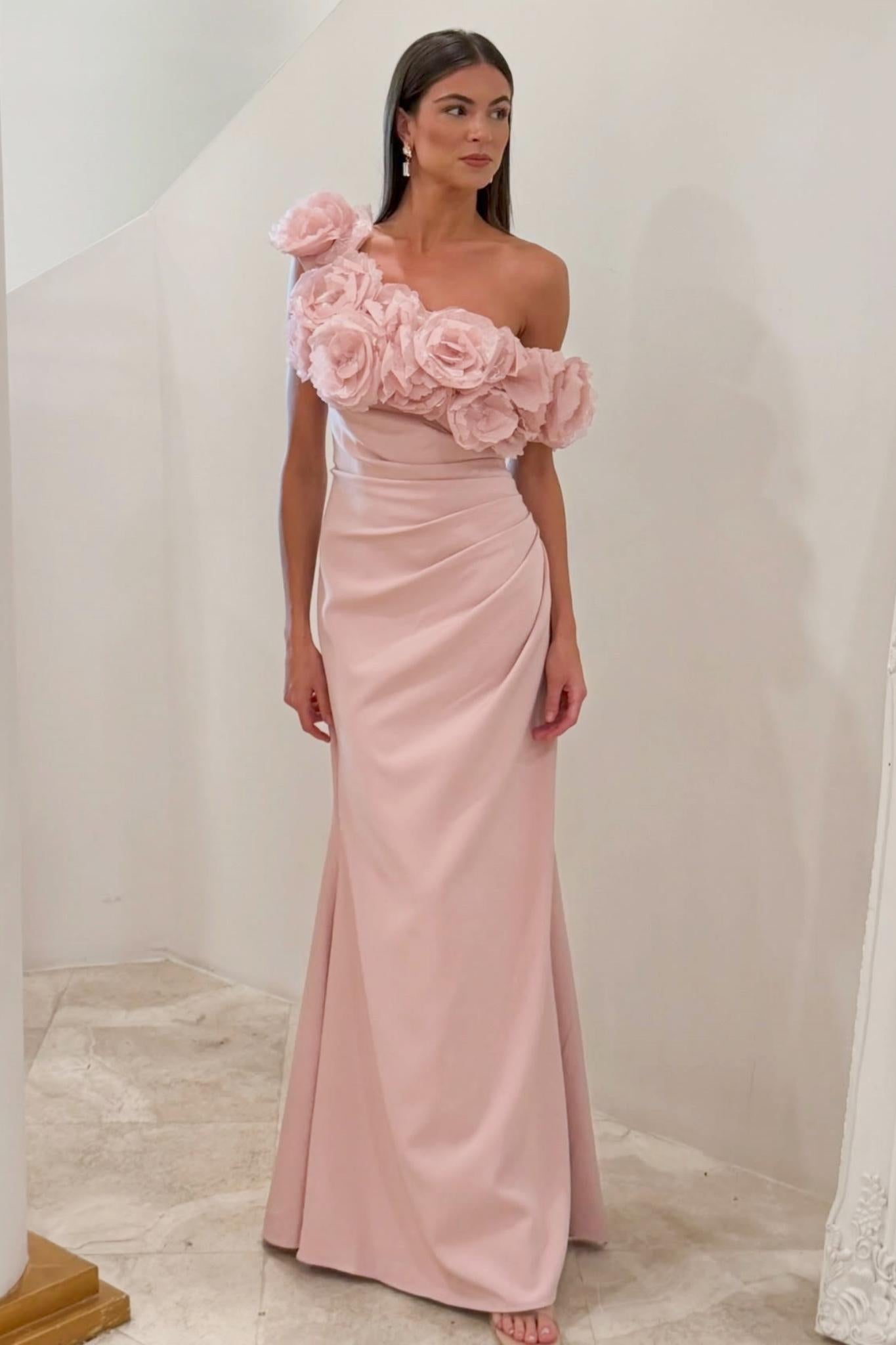 A woman modeling a floor-length blush pink gown with an asymmetric one-shoulder design, featuring large 3D fabric roses across the bodice and a draped skirt.