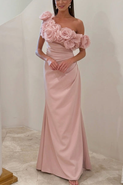 A woman modeling a floor-length blush pink gown with an asymmetric one-shoulder design, featuring large 3D fabric roses across the bodice and a draped skirt.