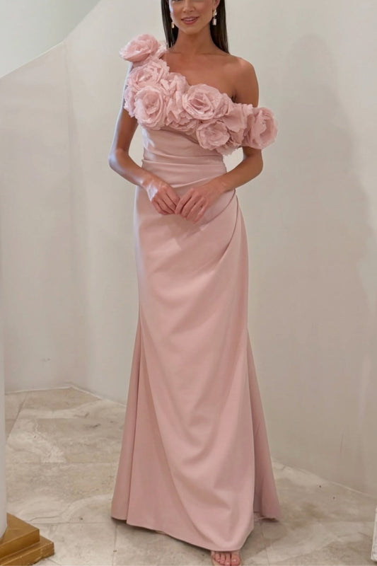 A woman wearing a floor-length one-shoulder formal gown in dusty pink. The dress features a large, structured floral applique on the off-shoulder neckline and a draped satin skirt, shown against a neutral indoor background.