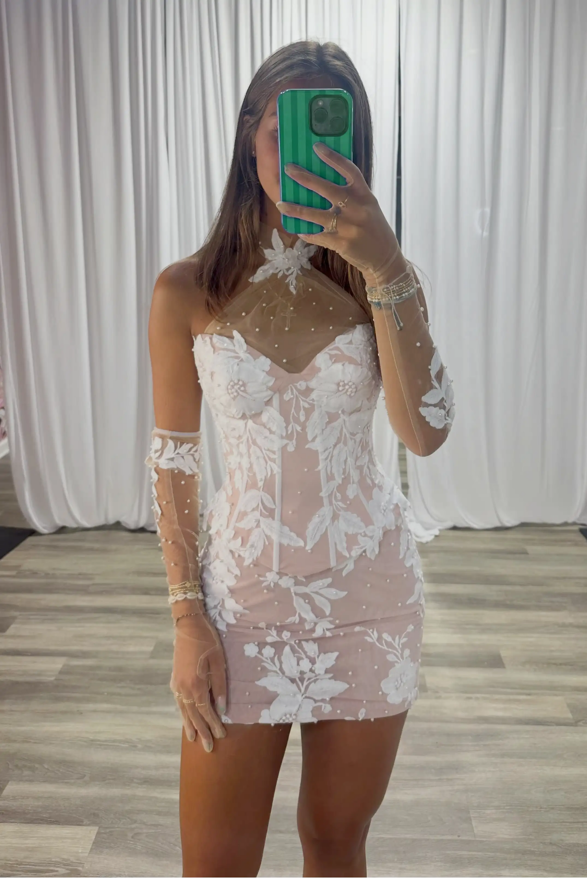 A woman takes a mirror selfie wearing a sheer blush pink mini dress with white floral lace patterns and long sheer sleeves. The background features white draped curtains and a light wood floor.