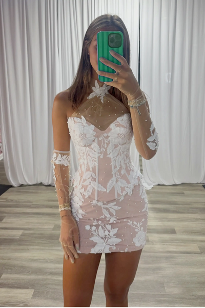 A woman takes a mirror selfie wearing a sheer blush pink mini dress with white floral lace patterns and long sheer sleeves. The background features white draped curtains and a light wood floor.