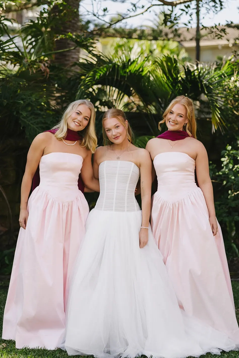 Three women wearing formal dresses pose together outdoors in front of lush green foliage. The two women on either side wear matching strapless blush pink floor-length gowns with deep maroon neck wraps.