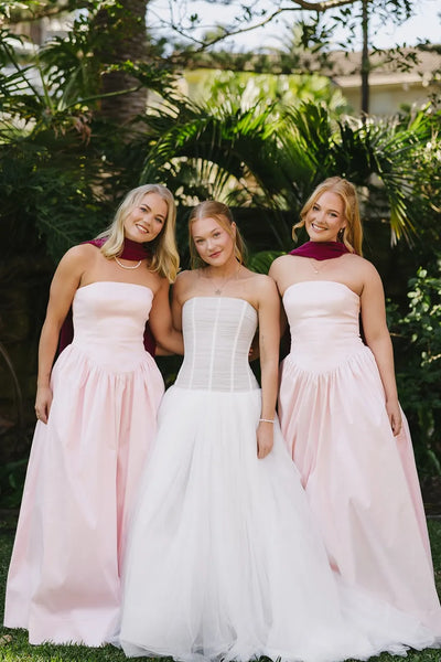 Three women wearing formal dresses pose together outdoors in front of lush green foliage. The two women on either side wear matching strapless blush pink floor-length gowns with deep maroon neck wraps.