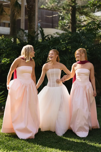 Three women wearing formal dresses pose together outdoors in front of lush green foliage. The two women on either side wear matching strapless blush pink floor-length gowns with deep maroon neck wraps.