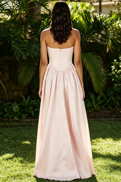 Three women wearing formal dresses pose together outdoors in front of lush green foliage. The two women on either side wear matching strapless blush pink floor-length gowns with deep maroon neck wraps.