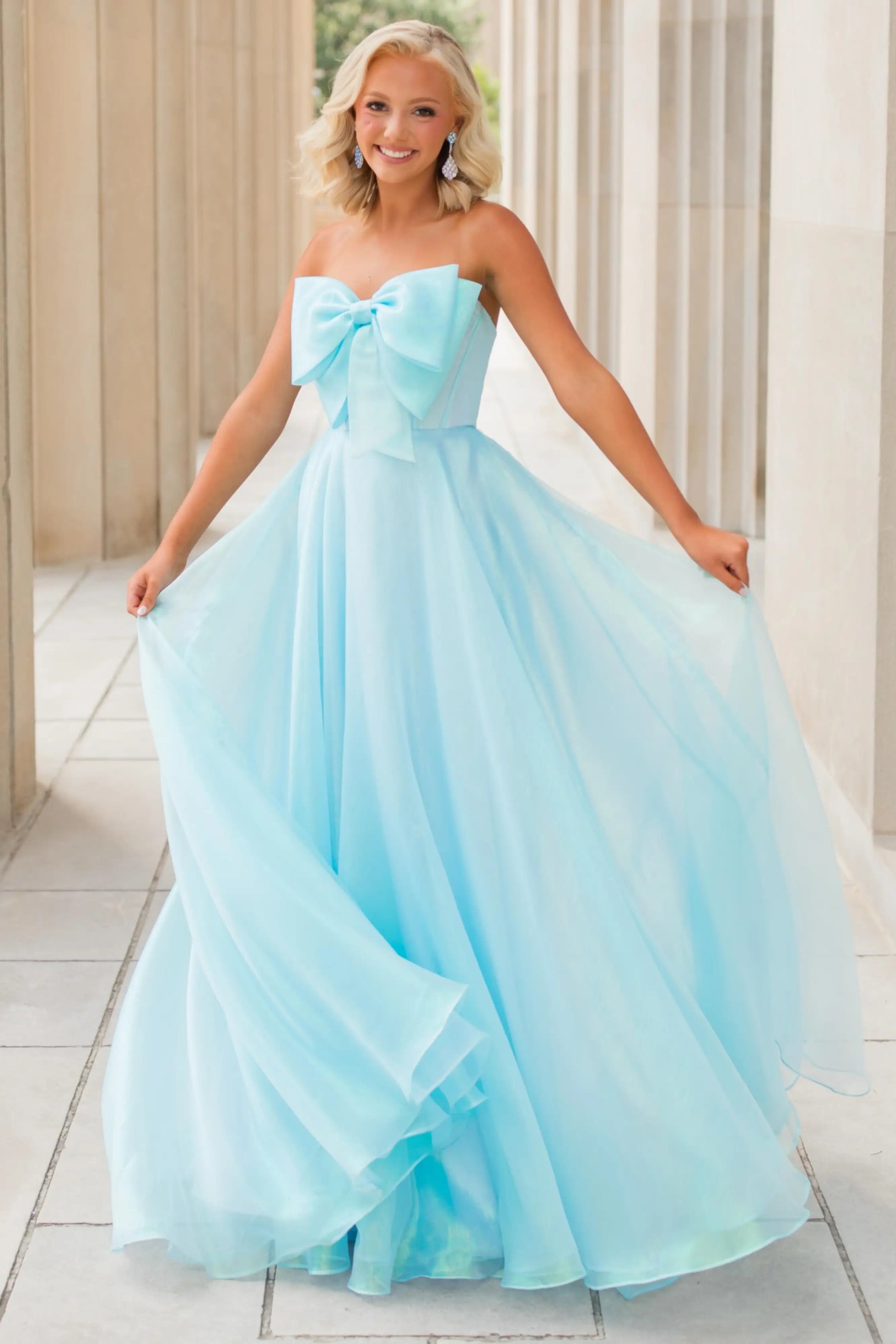 A young woman wearing a strapless light blue tulle prom dress with a large bow at the bust and a full flowing skirt, standing in a covered walkway.