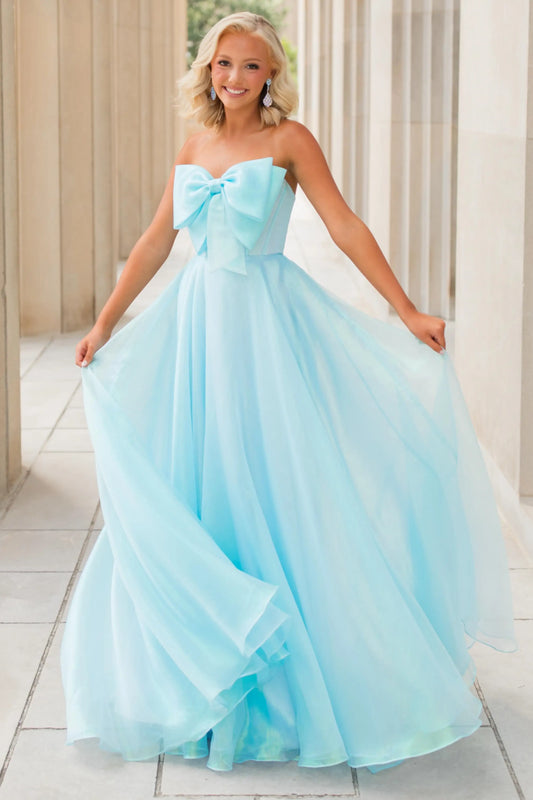 A young woman wearing a strapless light blue tulle prom dress with a large bow at the bust and a full flowing skirt, standing in a covered walkway.