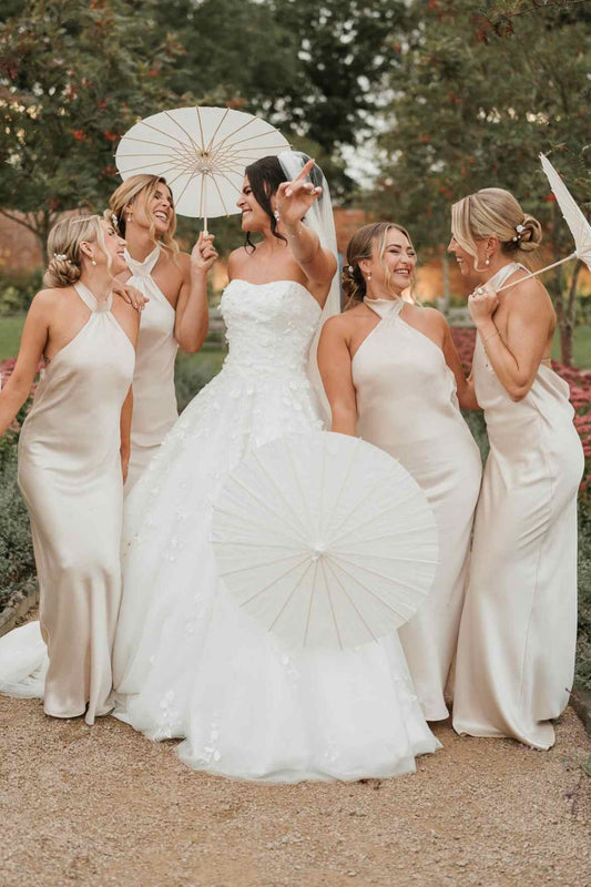 A bride in a strapless white wedding gown stands outdoors with four bridesmaids wearing matching ivory satin dresses, holding white paper umbrellas in a garden setting.