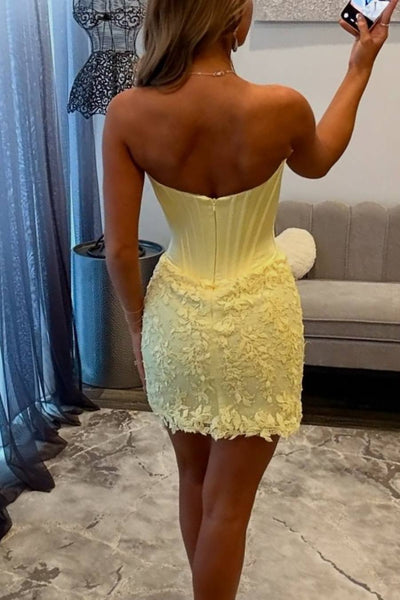 A woman takes a mirror selfie in a room wearing a yellow strapless homecoming dress with a corset-style bodice, paired with white high-heeled sandals.