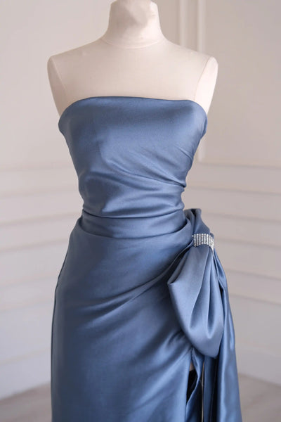 A strapless floor-length satin gown in a dusty slate blue color, displayed on a cream mannequin in an elegant indoor setting with white paneled walls and light wood flooring.
