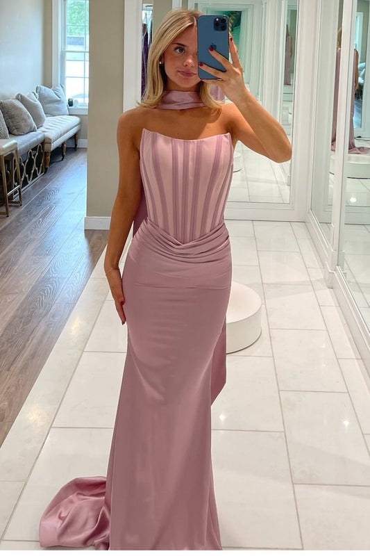 A woman takes a mirror selfie in a fitting room wearing a floor-length dusty pink gown with a strapless sweetheart bodice, vertical pleats, and a draped neck detail.