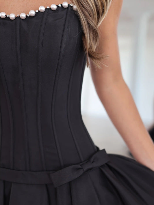 Close-up of a black sleeveless dress with a corset-style bodice, pearl embellishment along the neckline, and a matching waist tie, worn by a model.