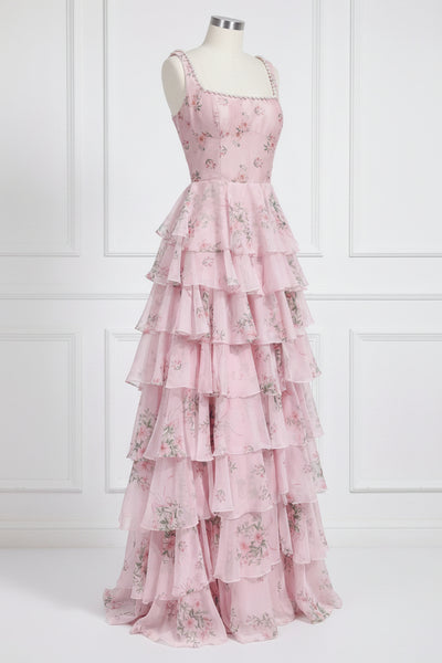 A pink tiered maxi dress with a floral print, displayed on a mannequin against a white paneled wall.