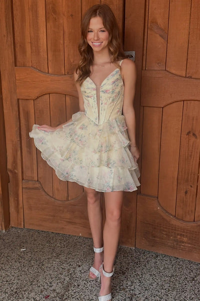 A woman modeling a short, ruffled spaghetti-strap dress with a corset-style bodice and a light floral print, standing in front of a rustic wooden door.