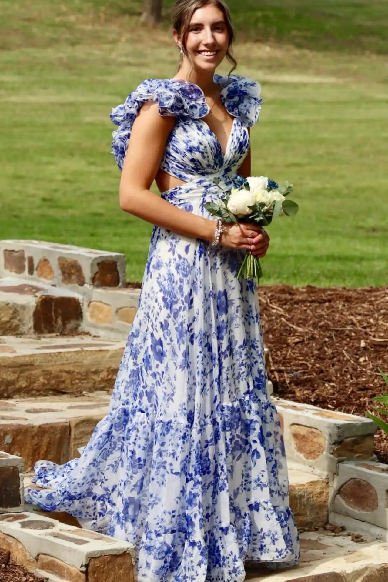 A woman wearing a blue and white floral print maxi dress with ruffled cap sleeves and a side cutout, holding a bouquet of white flowers while standing on outdoor stone steps.