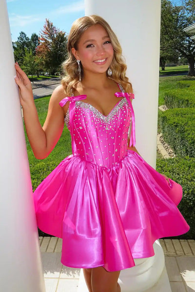 A woman modeling a short, bright pink satin homecoming with a sweetheart neckline, embellished bodice, and flared skirt, standing outdoors on brick steps in front of a house.