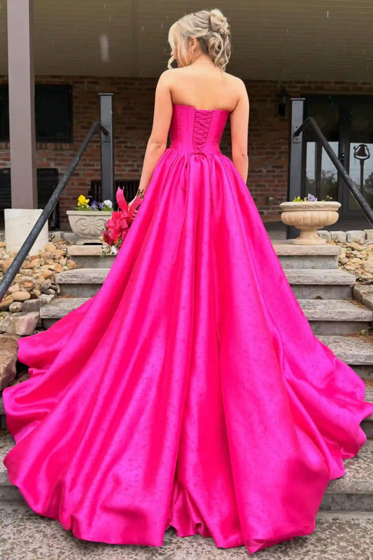A woman viewed from behind wearing a bright hot pink strapless ball gown with a corset-style back and a full satin-like skirt, standing on outdoor stone steps in front of a building.