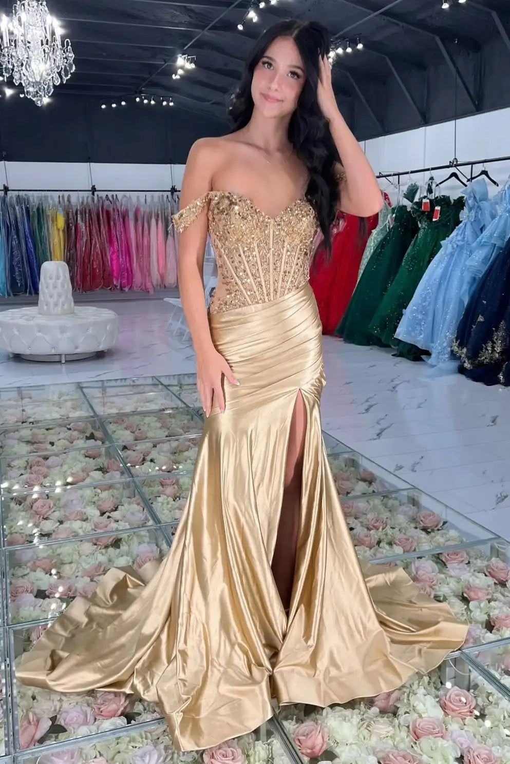 A woman models an off-the-shoulder  gold mermaid gown with a sweetheart neckline, heavily embellished bodice, and a high thigh slit, set in a dress showroom.
