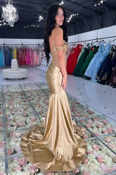A woman models an off-the-shoulder  gold mermaid gown with a sweetheart neckline, heavily embellished bodice, and a high thigh slit, set in a dress showroom.
