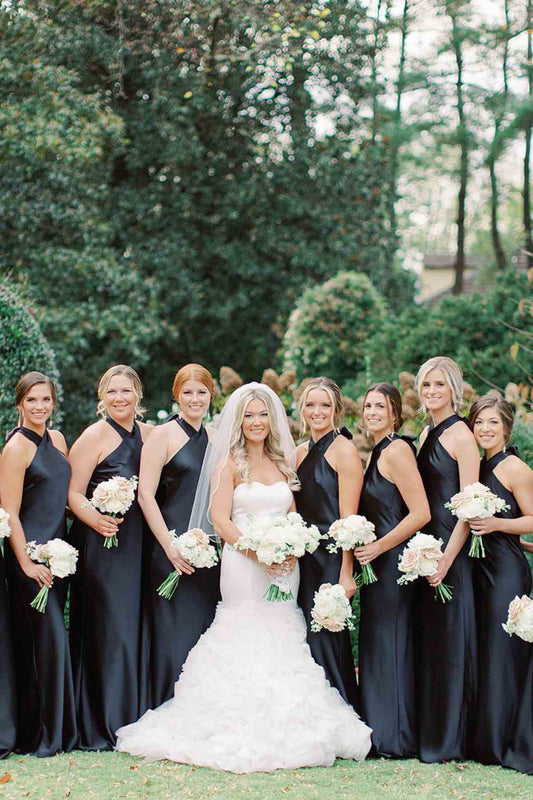 A bridal party posing outdoors in front of lush green trees. The bride is in the center wearing a white tulle wedding gown and veil, holding a bouquet of white flowers.