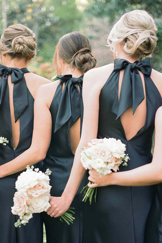 Three bridesmaids seen from behind wearing matching black halter-neck dresses with large bow back details, holding white and blush rose bouquets outdoors.