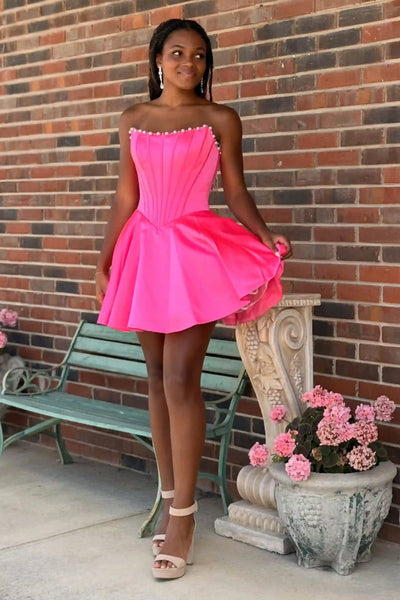 A woman wearing a bright hot pink strapless mini dress with a corseted bodice and flared skirt, standing outdoors in front of a brick wall next to a green bench and a planter.