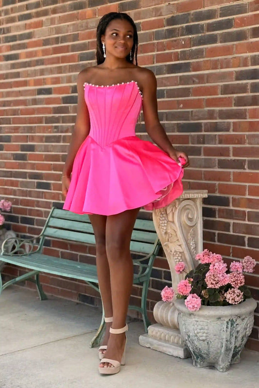 A woman modeling a bright hot pink strapless pleated mini dress with a flared skirt, standing outdoors against a red brick wall next to a green bench and a stone planter with pink flowers.