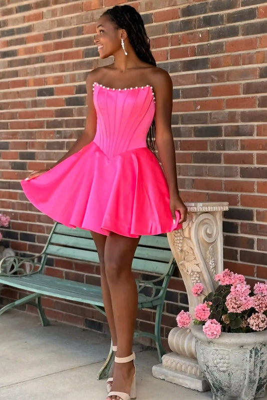 A woman wearing a bright hot pink strapless mini dress with a corseted bodice and flared skirt, standing outdoors in front of a brick wall next to a green bench and a planter.