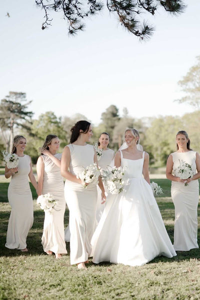 Five women wearing matching sleeveless floor-length bridesmaid dresses in a soft color, standing together in a decorated room with green drapes.
