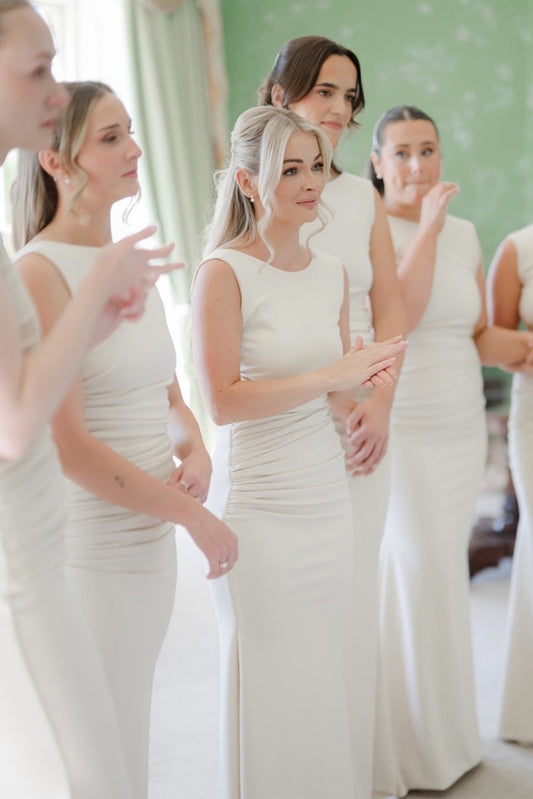 A group of women wearing matching fitted sleeveless dresses in a soft ivory tone, standing indoors in a bright room with a pale green wall and light curtains.