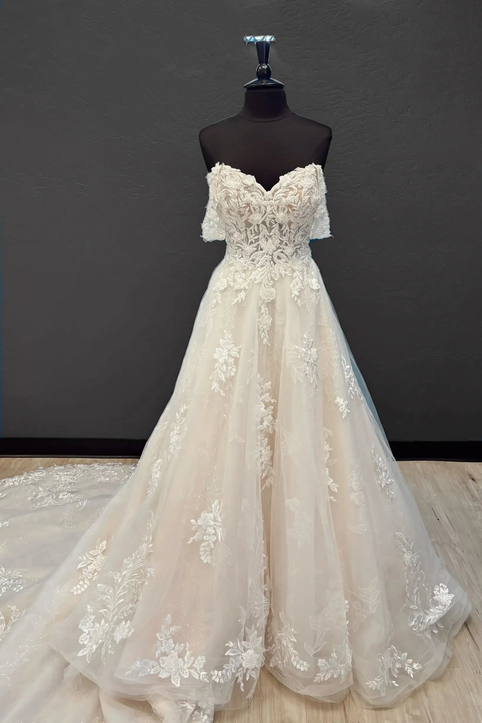 An off-the-shoulder A-line wedding gown in champagne tulle with intricate floral lace appliques, displayed on a black mannequin dress form against a dark gray wall.