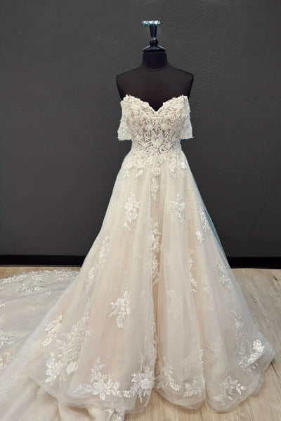An off-the-shoulder A-line wedding gown in champagne tulle with intricate floral lace appliques, displayed on a black mannequin dress form against a dark gray wall.