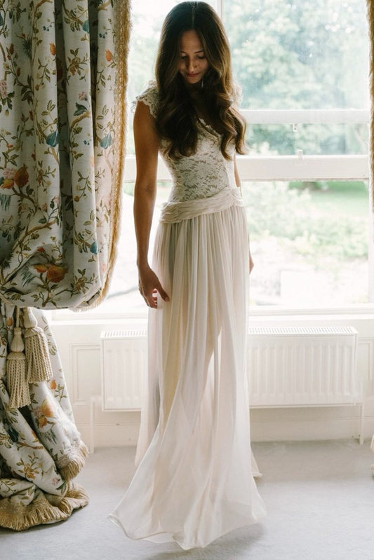 A woman wearing a sleeveless cream maxi dress with a lace bodice and flowing tulle skirt, standing in a bright room with a window and floral curtains.