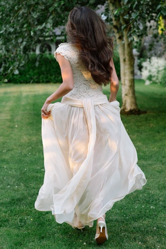 A woman wearing a cream-colored lace and tulle dress is captured from behind as she walks on a green lawn outdoors.