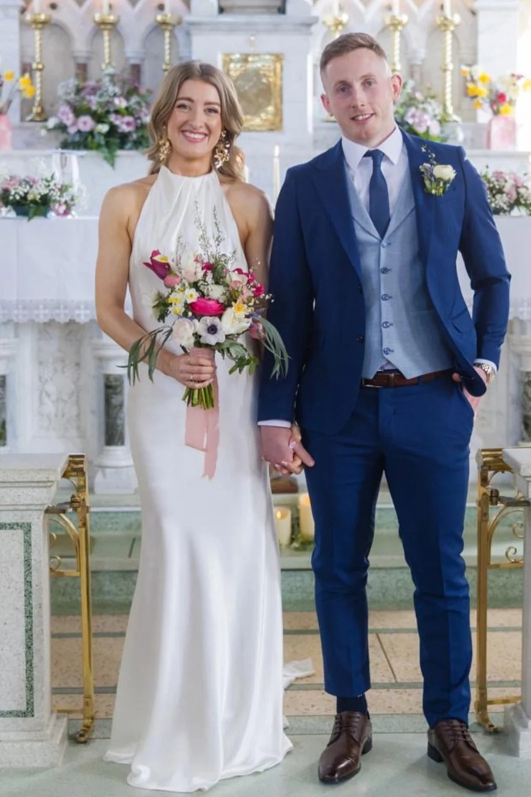 A bride in a sleeveless ivory halter-neck wedding gown holding a colorful bouquet and standing next to a groom in a navy blue three-piece suit with a light blue vest and tie inside a church setting.