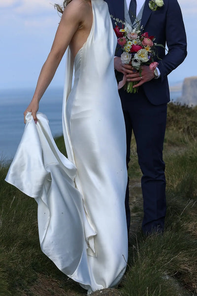 A bride in a sleeveless ivory halter-neck wedding gown holding a colorful bouquet and standing next to a groom in a navy blue three-piece suit with a light blue vest and tie inside a church setting.