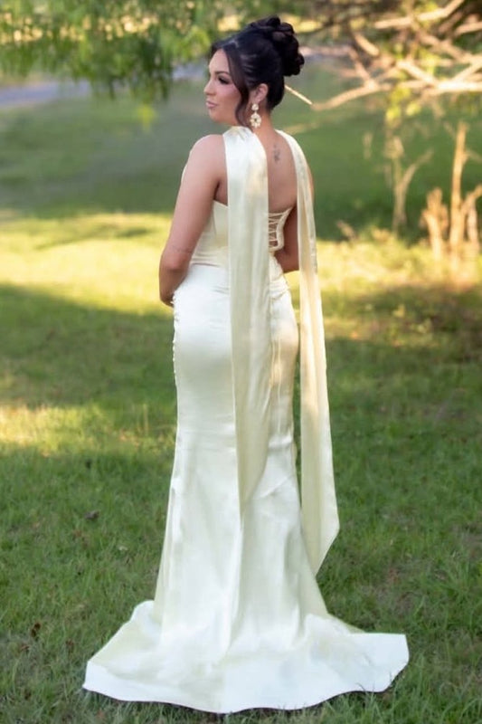 A woman stands outdoors on grass wearing a fitted ivory prom dress with a long draped train and backless open back. 