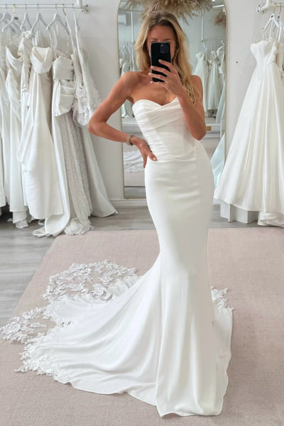 A woman takes a mirror selfie in a bridal shop wearing a strapless ivory mermaid-style wedding gown with a sweetheart neckline, fitted bodice, and a long lace-trimmed train. White wedding dresses are hanging in the background.