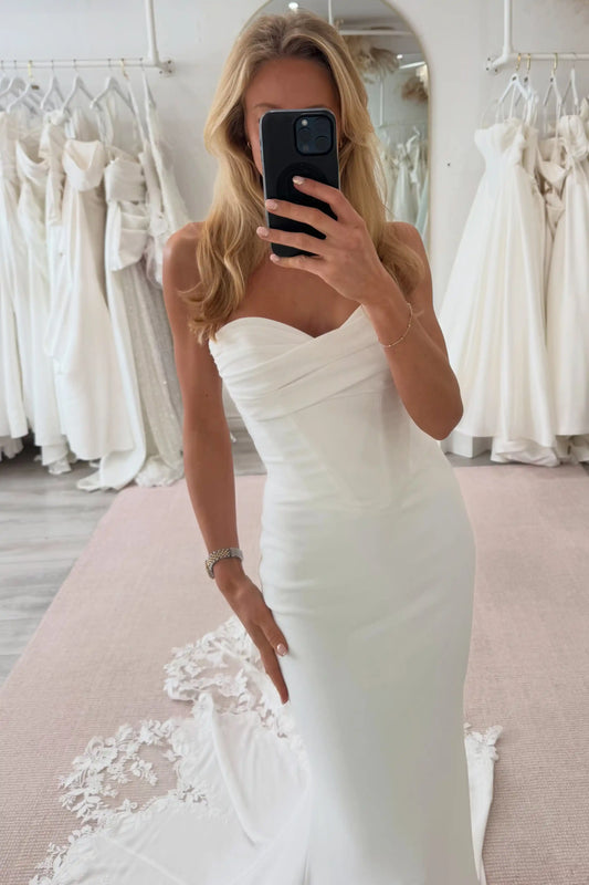 A woman takes a mirror selfie in a bridal shop wearing a strapless ivory wedding gown with ruching and a fitted silhouette. White wedding dresses are hanging in the background, and a pale pink carpet is visible beneath her.
