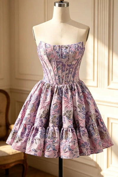 A short off-the-shoulder dress with a floral print in shades of pink, purple, and green, displayed on a mannequin in an indoor setting with warm sunlight and panelled walls.
